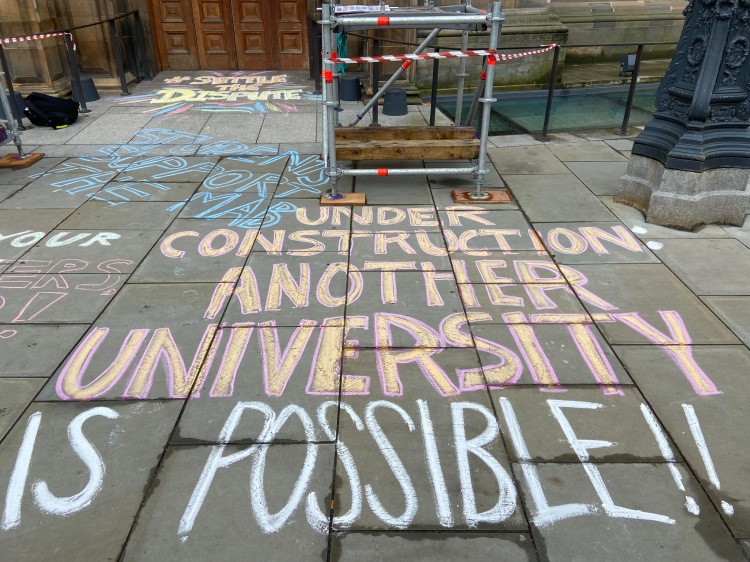 the words 'Under construction: another university is possible' chalked in large letters on a pavement in front of building and some scaffolding polls.
