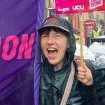 Sophia Woodman: Sophia is on a picket line holding a banner and a placard. She's wearing a cap and a cagoule.